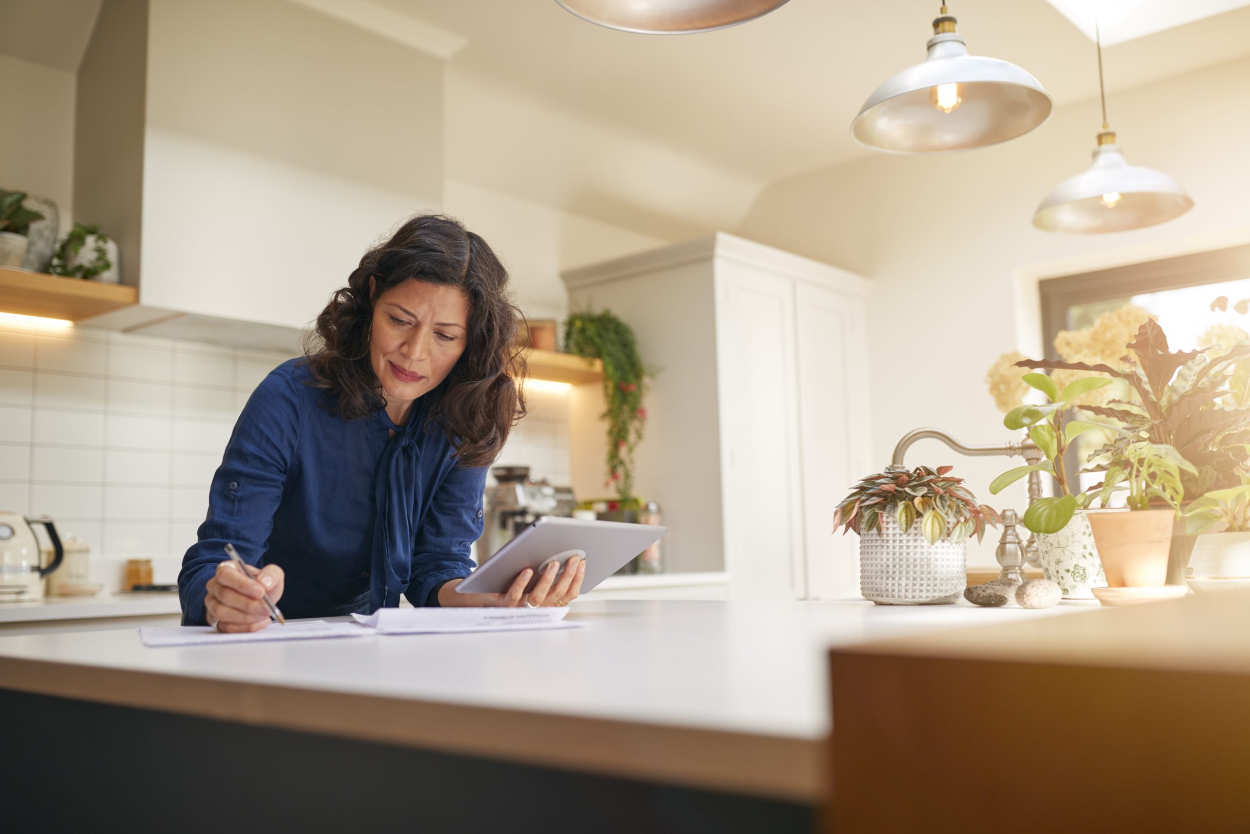 Mature Woman With Digital Device Reviewing Paperwork In Kitchen and thinking About who She will appoint as an executor of her will.